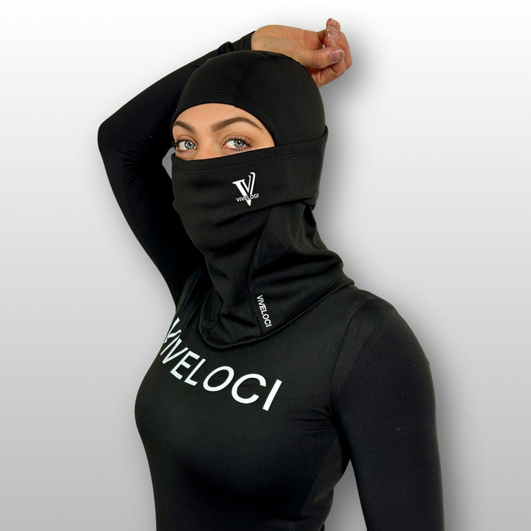 Bella wearing a black balaclava and black bodysuit with 'VIVELOCI' branding on a light gray background
