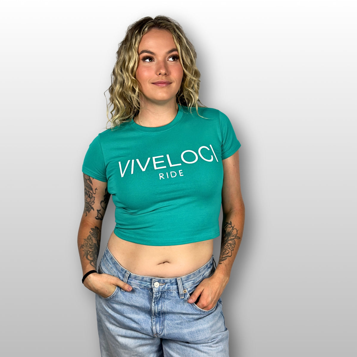 Emily is wearing a teal ViVeloci RIDE Crop top and blue jeans against a gray background