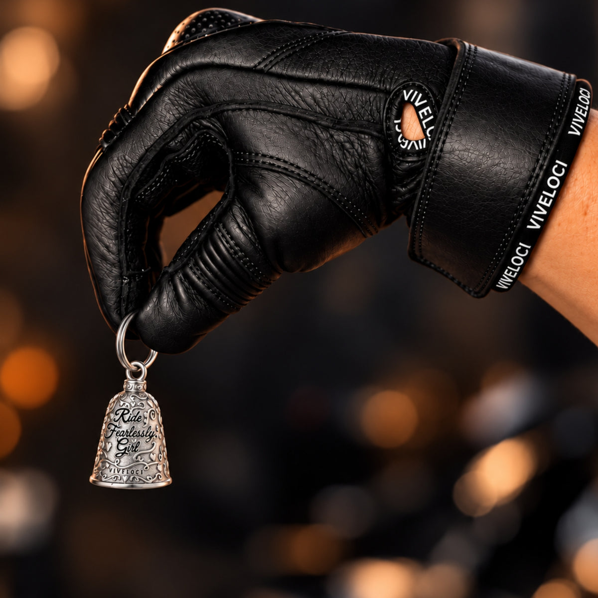 A close-up of a hand wearing a black leather motorcycle glove holding a small silver bell-shaped charm engraved with the words 'Ride Fearlessly, Girl'. The glove has 'VIVELOCI' printed on the wrist strap, and the background is a dark, blurred bokeh with warm golden lights.