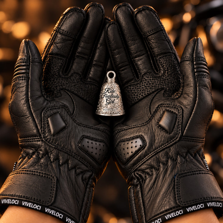 Black leather motorcycle gloves with 'VIVELOCI' branding on the wrist straps, held open to display a small silver bell-shaped pendant engraved with 'Ride Fearlessly, Girl'.