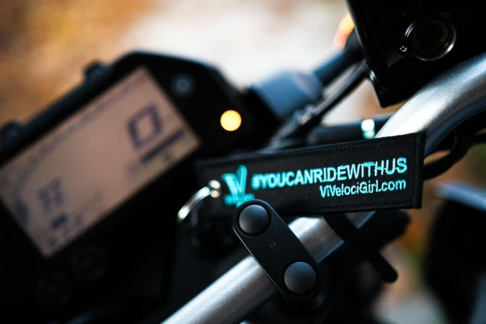 A close-up shot of a black motorcycle handlebar strap holding a keychain with a bright teal embroidery that reads '#YOUCANRIDEWITHUS' and 'VVelociGirl.com'.