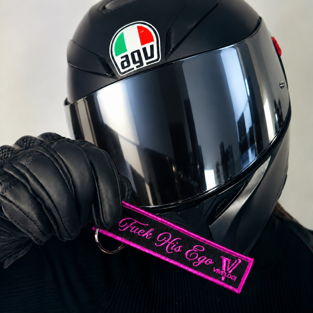 A close-up of a black AGV full-face motorcycle helmet with a dark tinted visor, held by a black leather glove. The glove is holding a black key tag with Fuck His Ego and the Viveloci logo embroidered in pink,