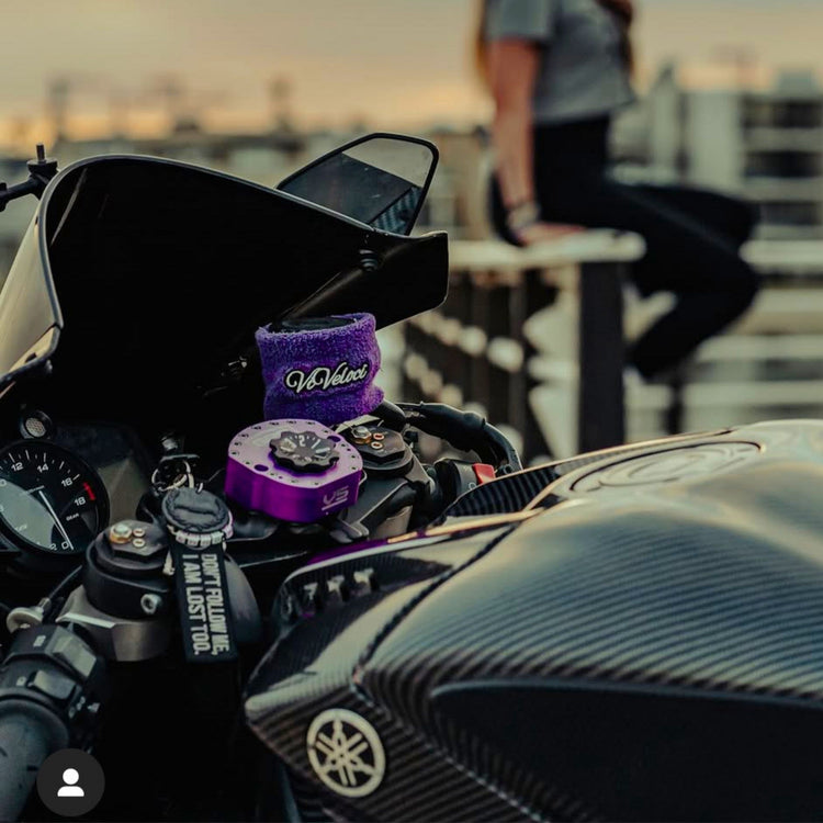 A close-up of a motorcycle with a purple ViVeloci reservoir cover set against a blurred background of a woman sitting on a railing at sunset.