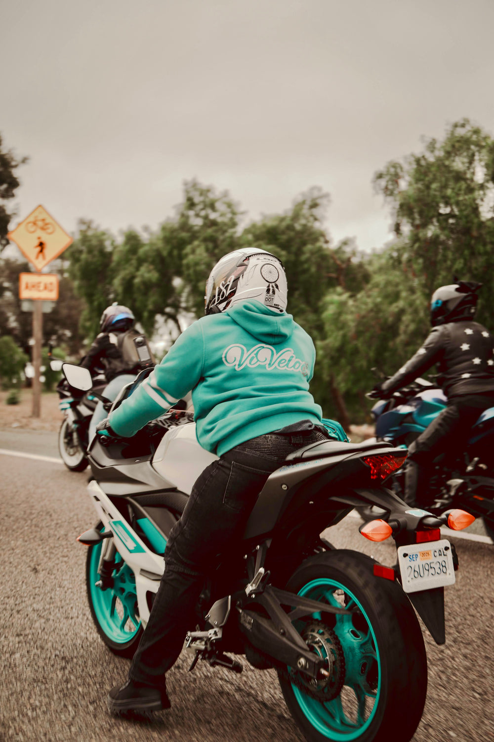 Woman on a motorcycle wearing a teal ViVeloci hoodie, riding on a road with trees and signs in the background.