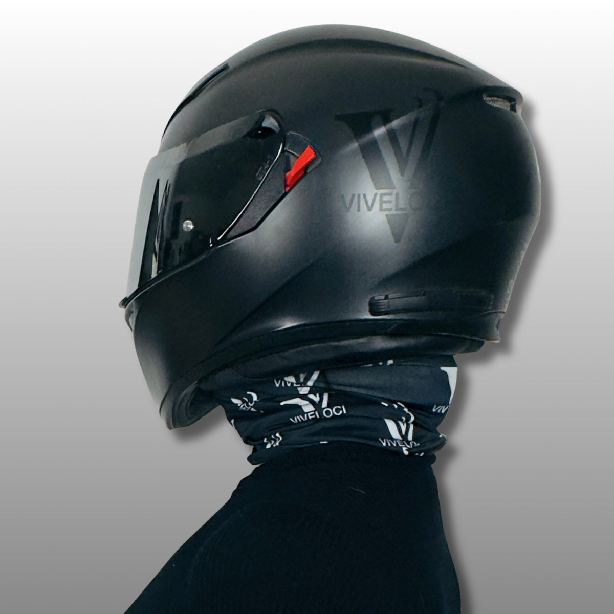 A matte black full-face motorcycle helmet, shown in side profile with a black ViVeloci Vinyl decal, on a person wearing a matching black neck gaiter with white VIVELOCI branding. The background is a clean, neutral gray gradient.